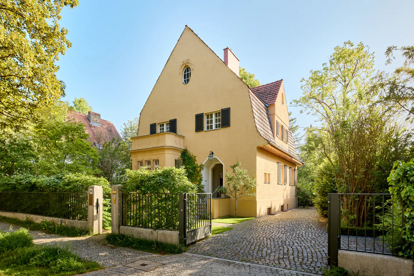 A yellow two-story house with a steep roof, black shutters, and a cobblestone driveway surrounded by lush green trees.