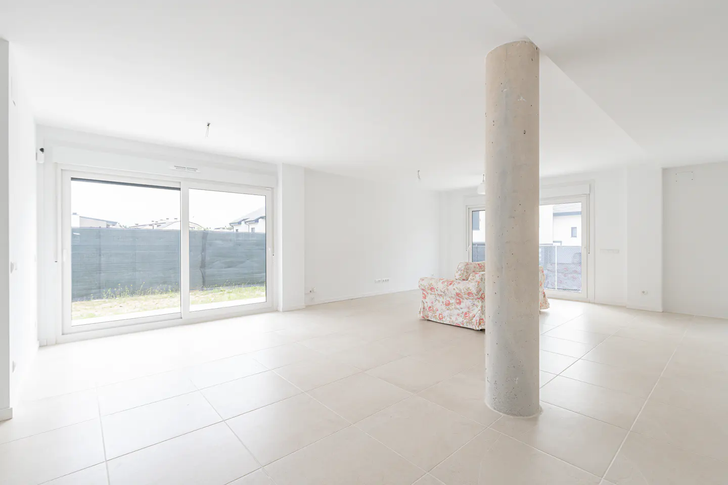 Bright, empty room with white walls, beige tile floor, and a concrete support column. A floral armchair sits near a window.