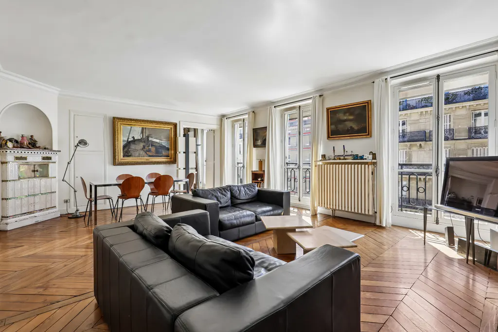Bright living room with parquet floors, black leather sofas, dining table, and windows overlooking a city view.