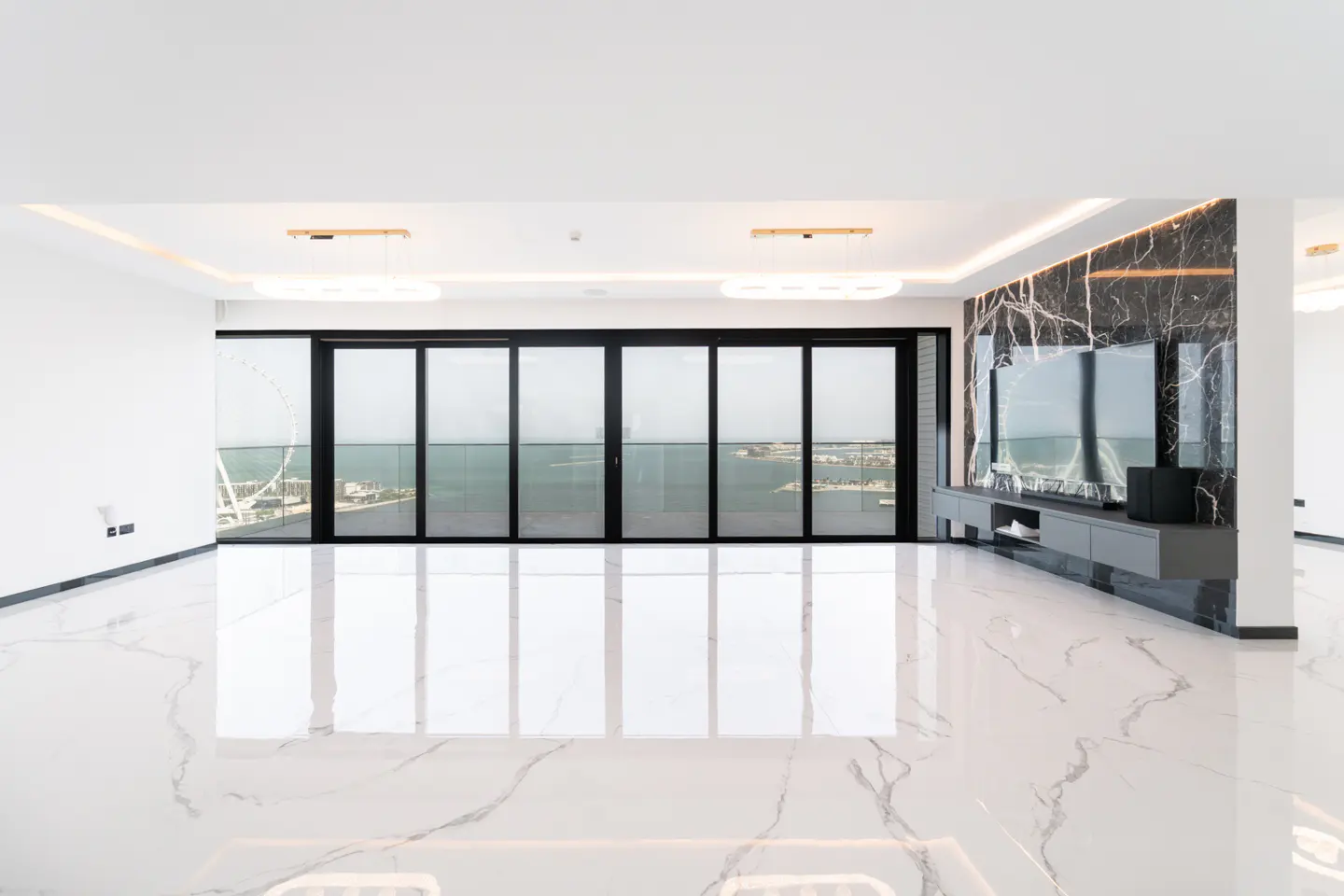Bright, empty room with white marble floors and a black marble accent wall with a TV. Black sliding glass doors lead to a balcony with an ocean view.