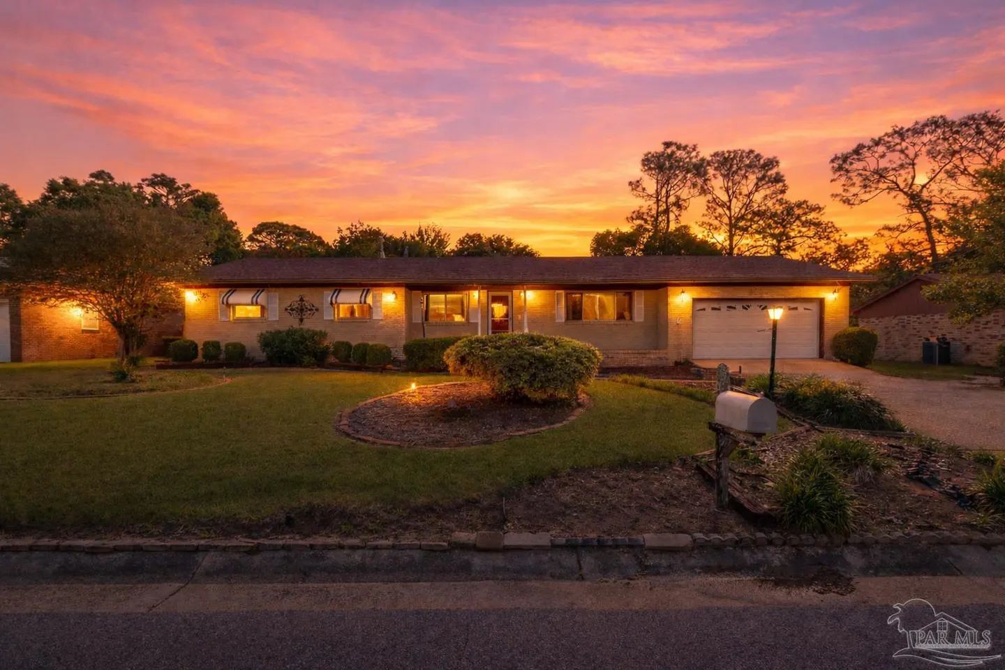 A ranch-style house with a green lawn at sunset. The sky is orange and pink. The house has a brown roof and white trim.