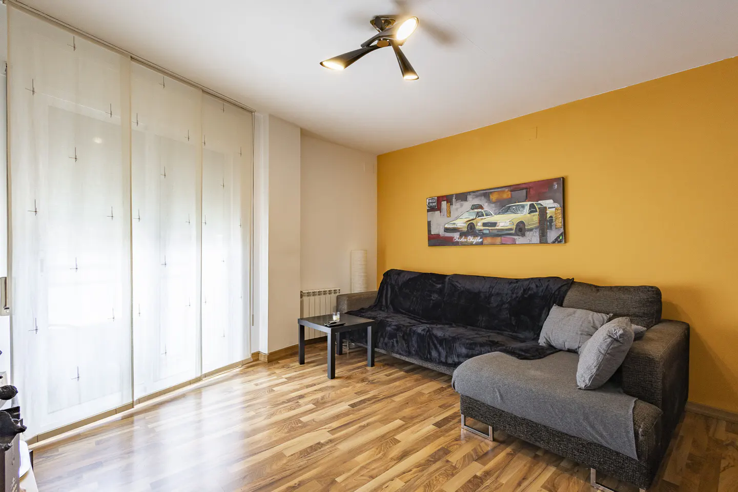 Living room with wood floors, yellow wall, and gray sofa with black throw. Car painting hangs above the sofa. Modern ceiling light.