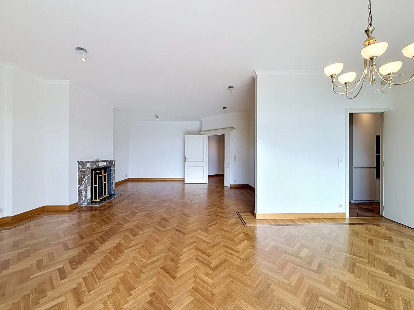 Spacious living room with white walls, herringbone wood floors, a marble fireplace, and a classic chandelier.