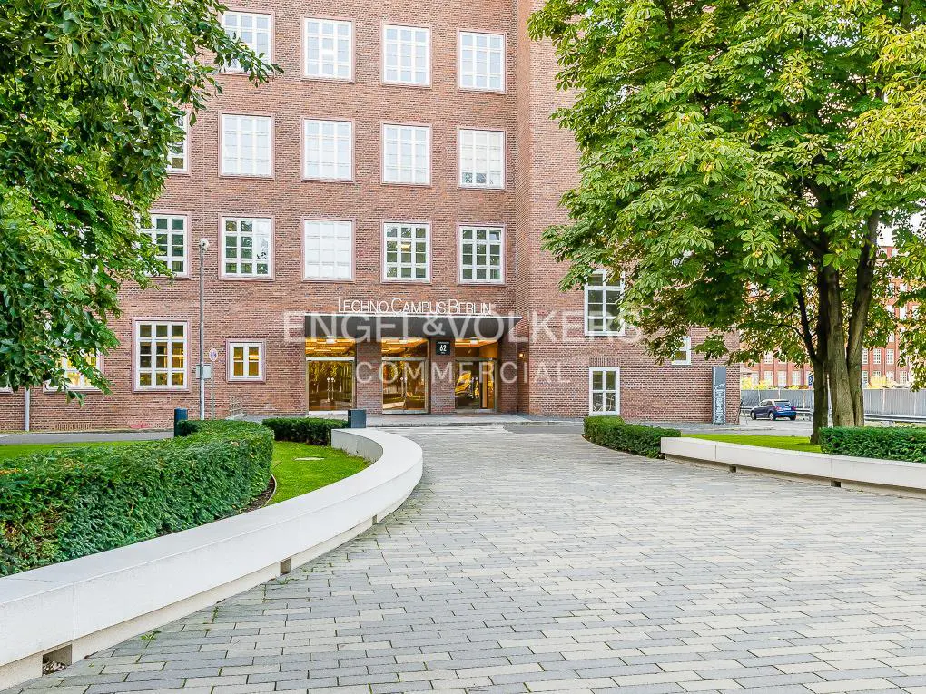 Exterior view of Engel & Völkers Commercial at Techno Campus Berlin. Brick building with white framed windows, trees, and a paved walkway.