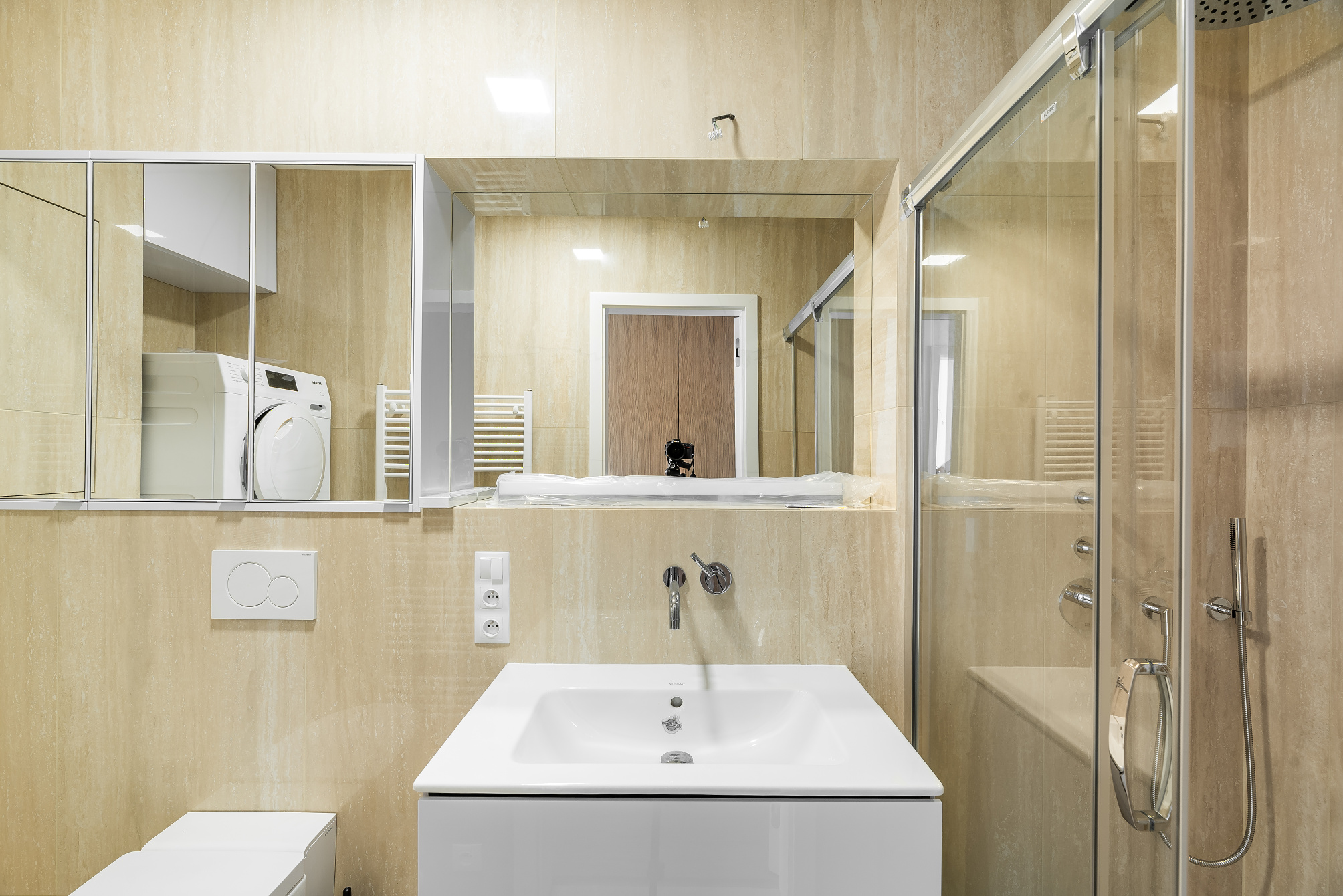 Modern bathroom with beige tiled walls, a white vanity sink, a glass shower enclosure, and a mirror reflecting a washer. [AI-generated text]