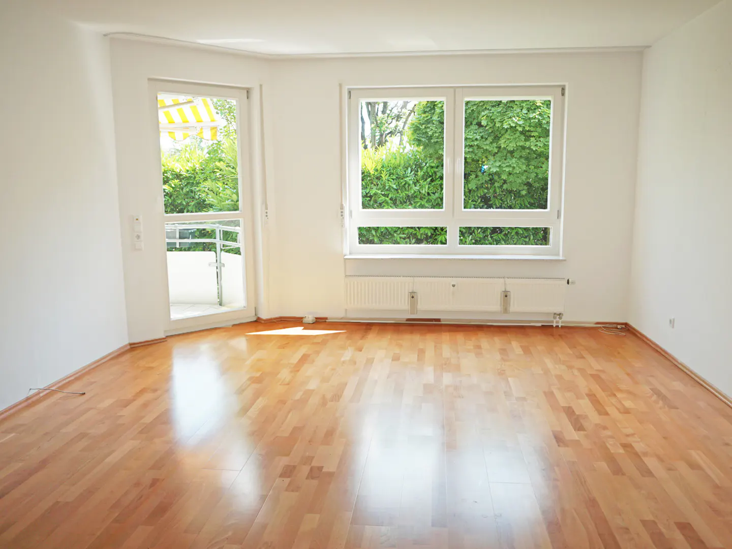 Bright, empty room with hardwood floors, white walls, a window overlooking green foliage, and a door to a balcony with a yellow awning.