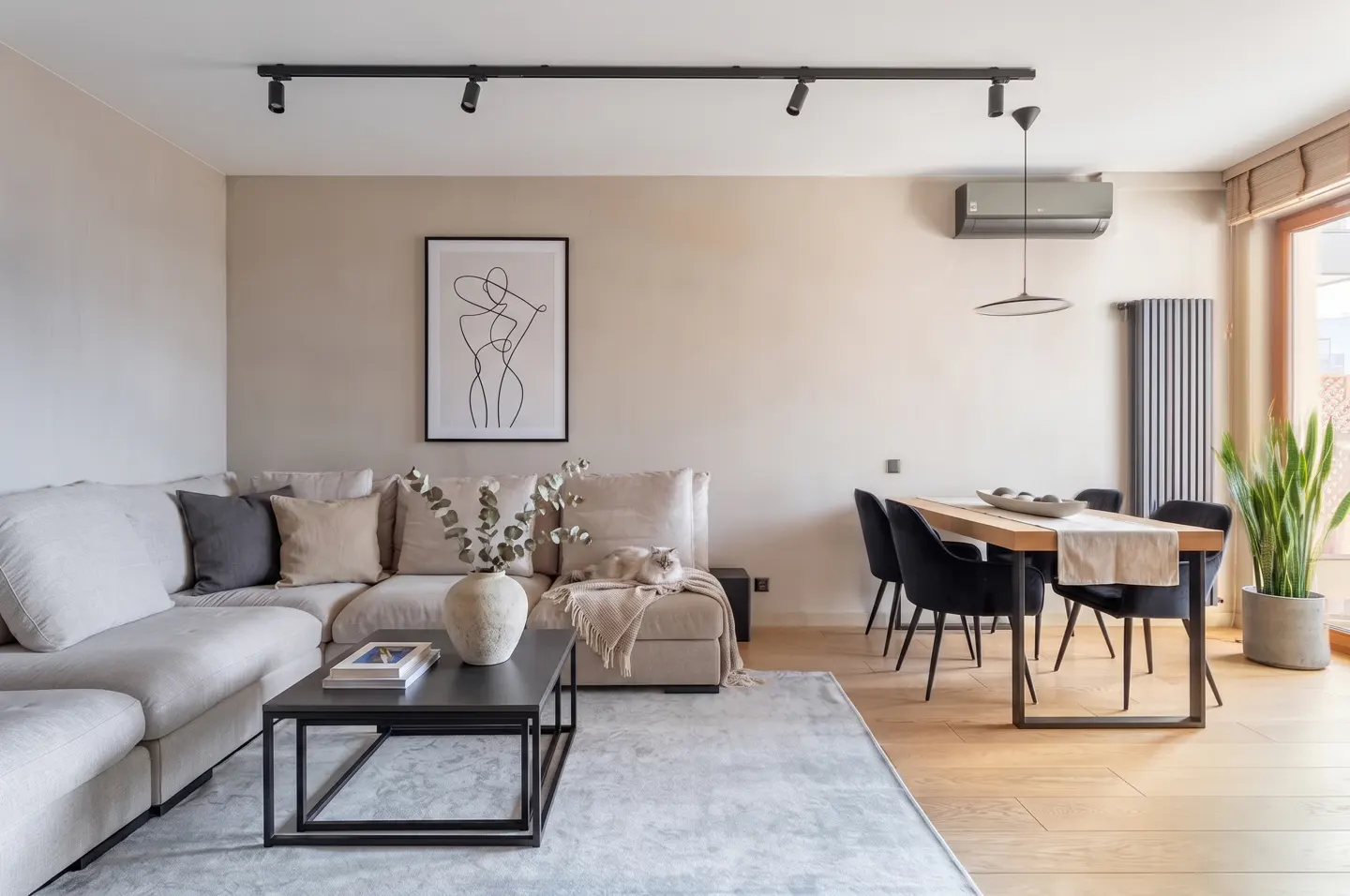 Living room with beige walls, a sectional sofa, black coffee table, and a dining table with black chairs. A cat is sleeping on the sofa.