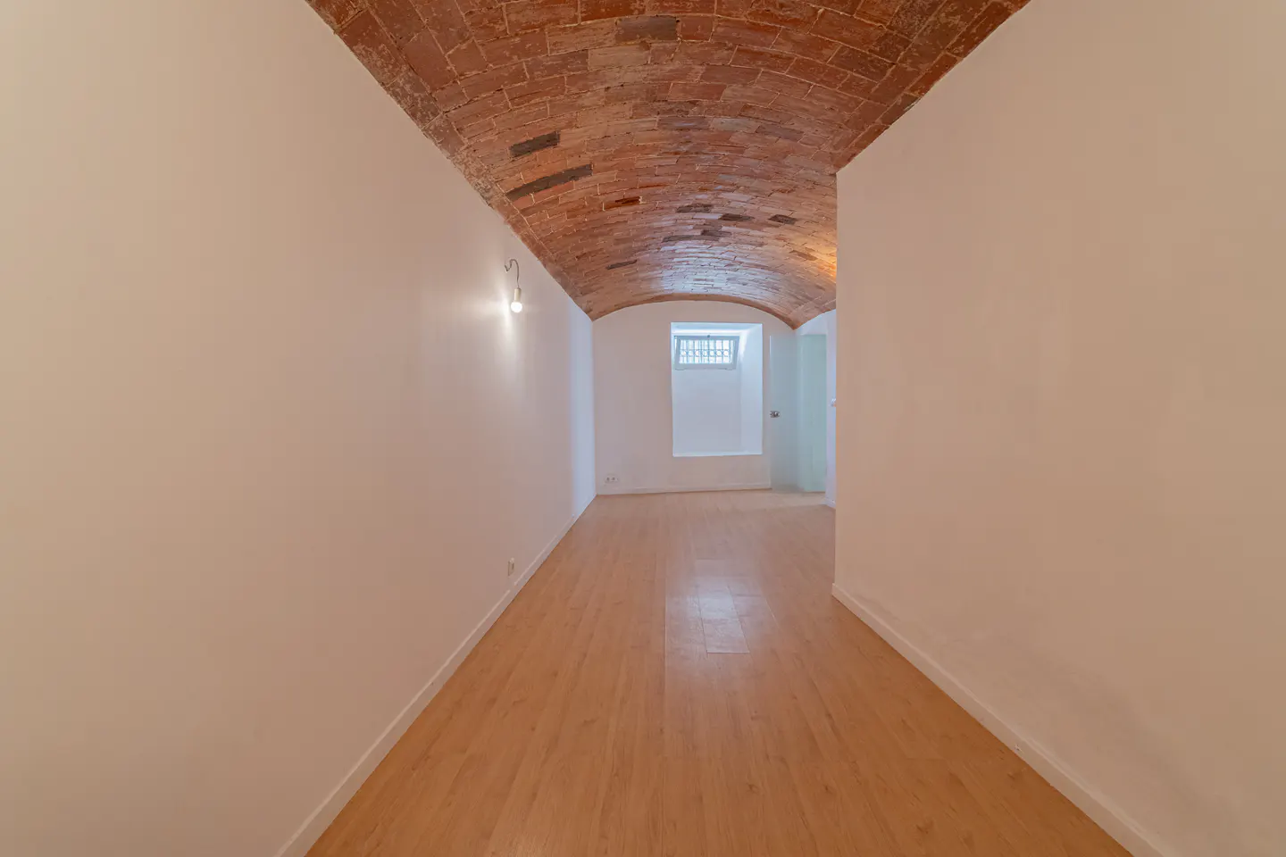 Empty room with light wood floors, white walls, and a brick arched ceiling. A window and doorway are visible at the end of the room.