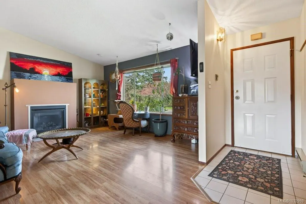 Living room with wood floors, fireplace, sunset painting, and large window with view of trees. White front door with floral rug.