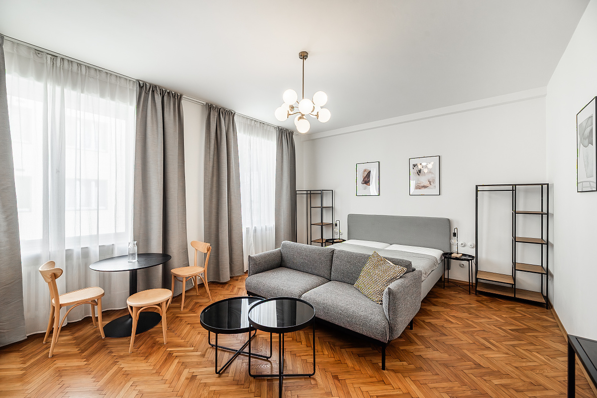 Modern studio apartment with a grey sofa, bed, two round coffee tables, dining set, and herringbone wood floors.
