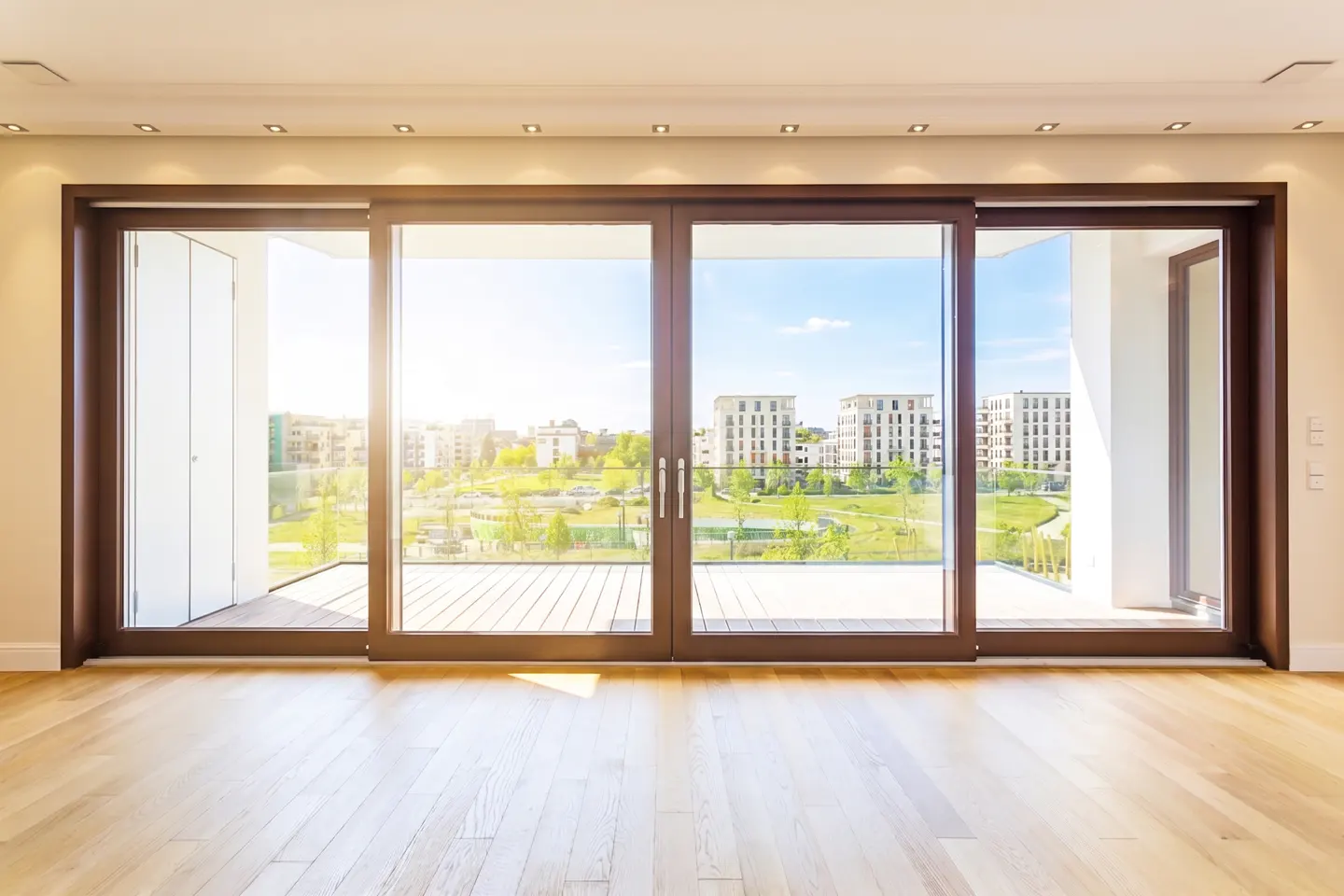 Bright room with wood floors and large sliding glass doors. Outside is a green space with buildings in the distance.