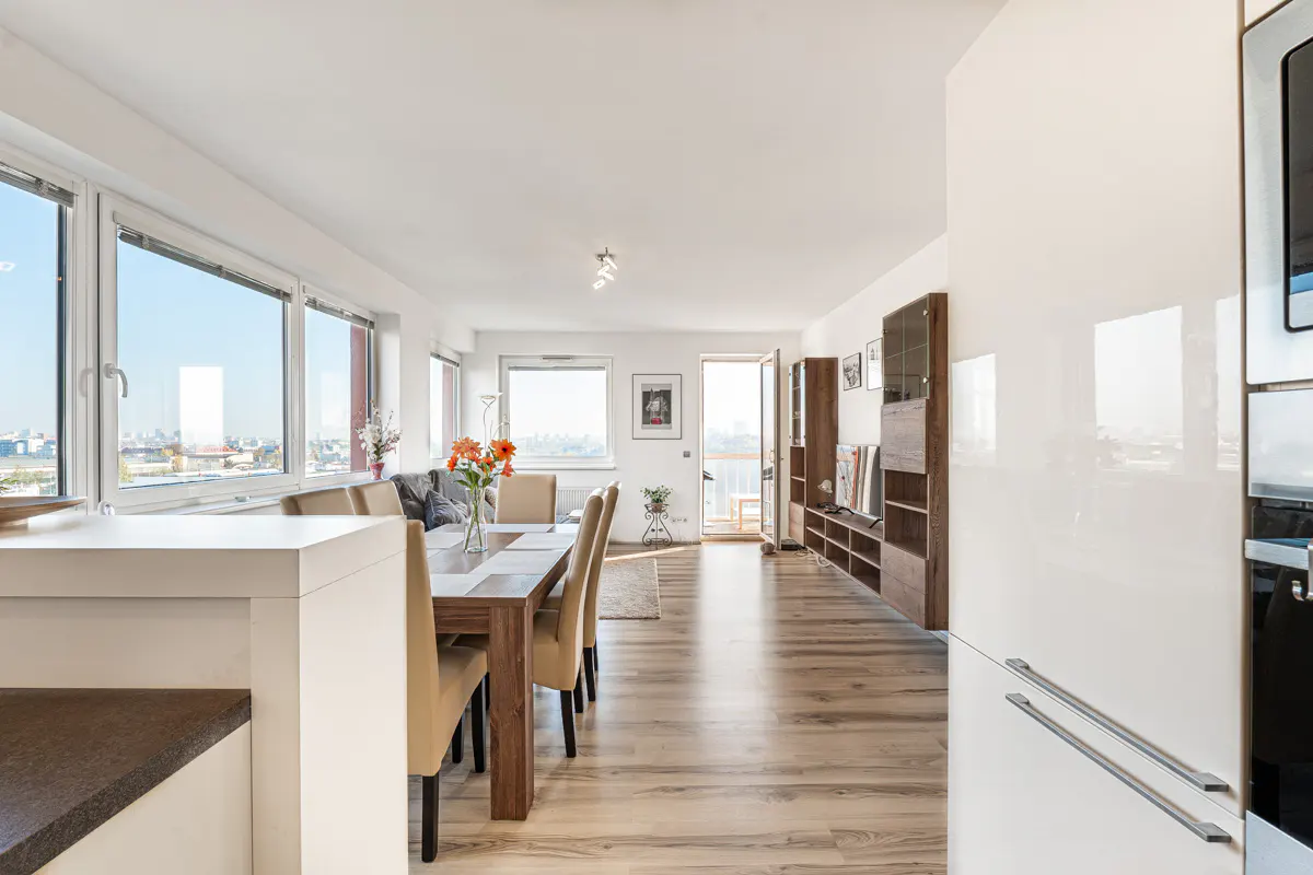 Bright open-plan living area with a wooden dining table, beige chairs, and large windows overlooking a city skyline.