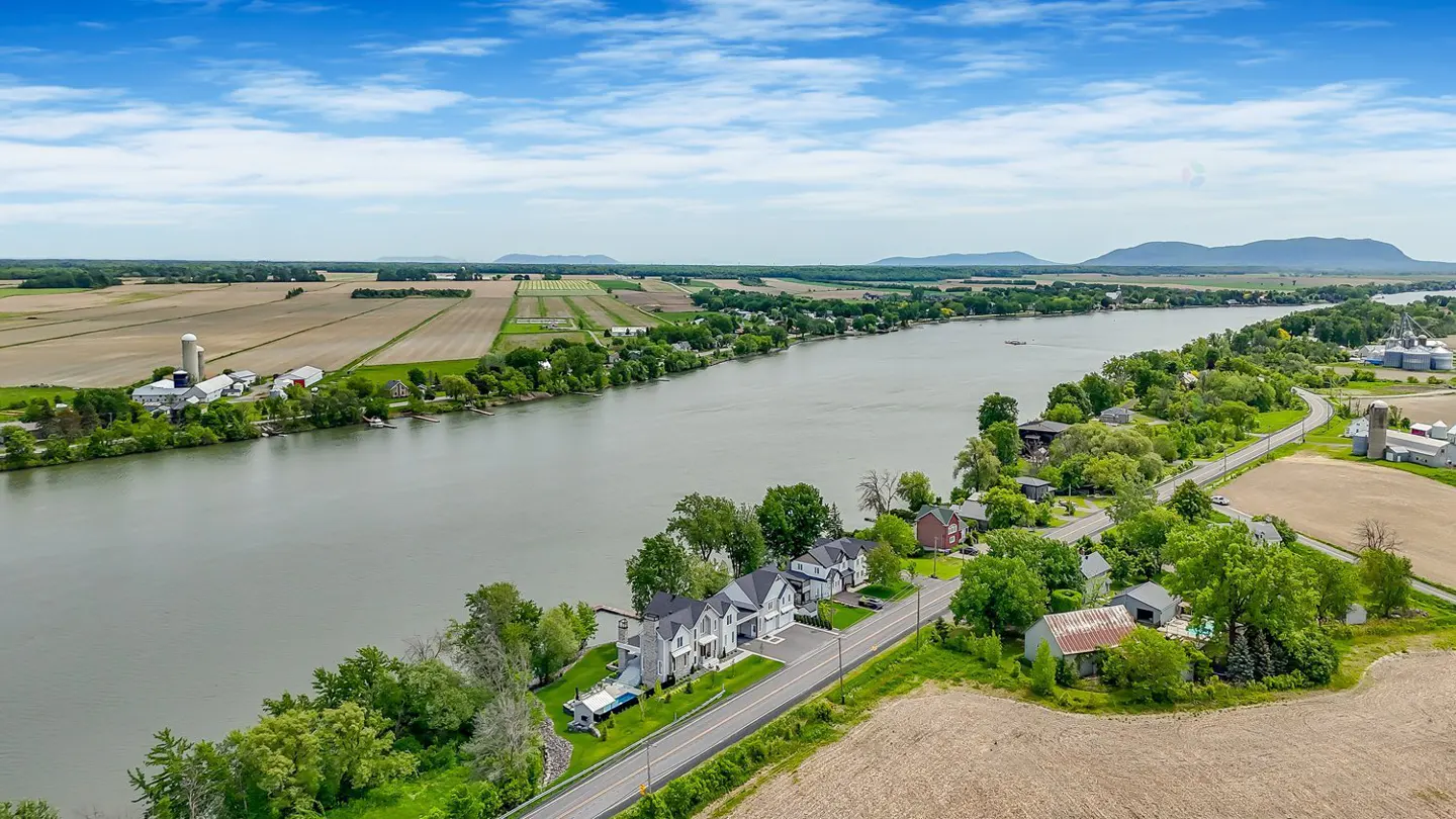 Aerial view of modern gray homes along a river, with farmland and mountains in the background under a blue sky.