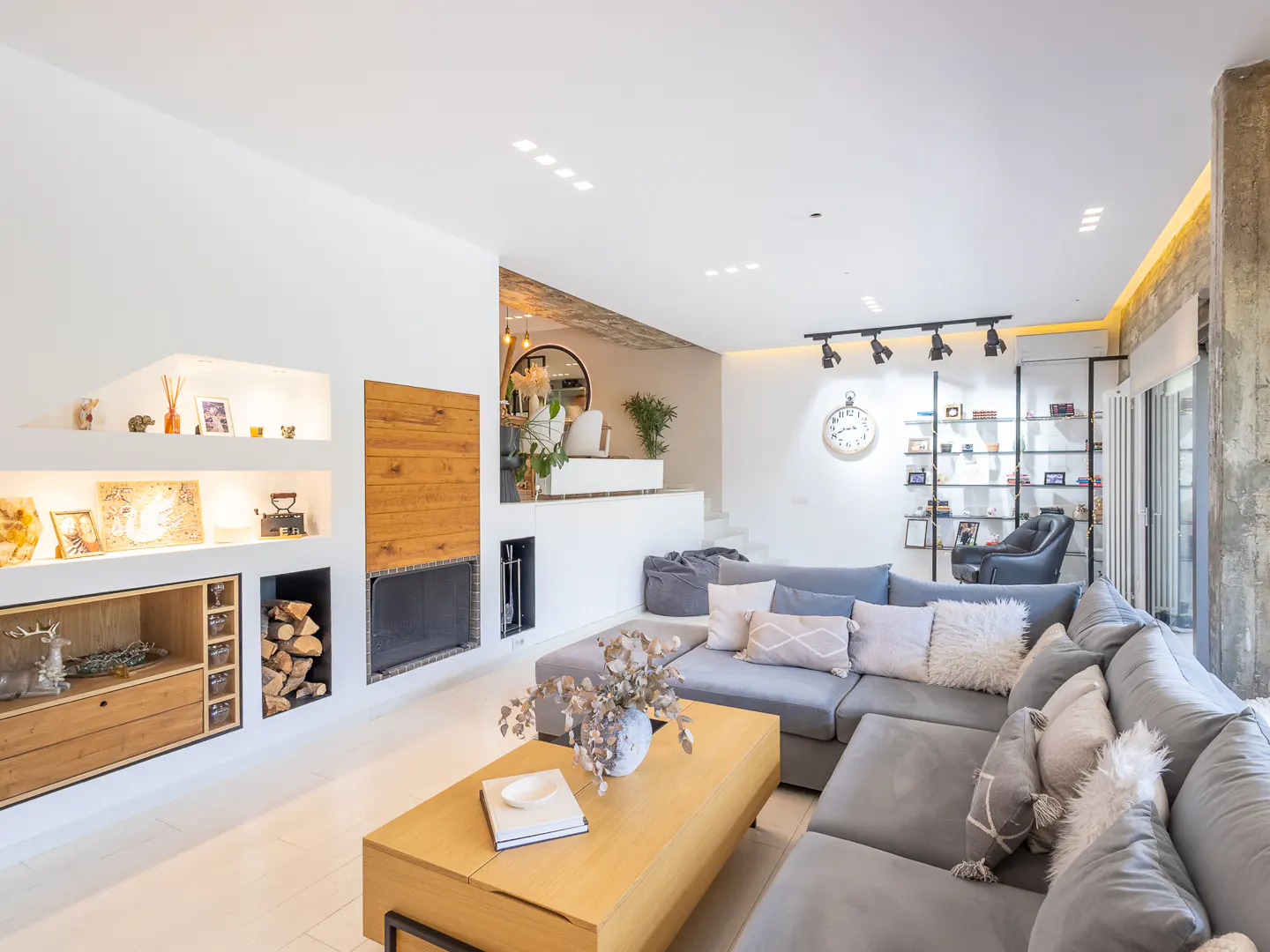 Modern living room with a grey sectional sofa, wooden coffee table, fireplace with firewood, and built-in wall shelving.