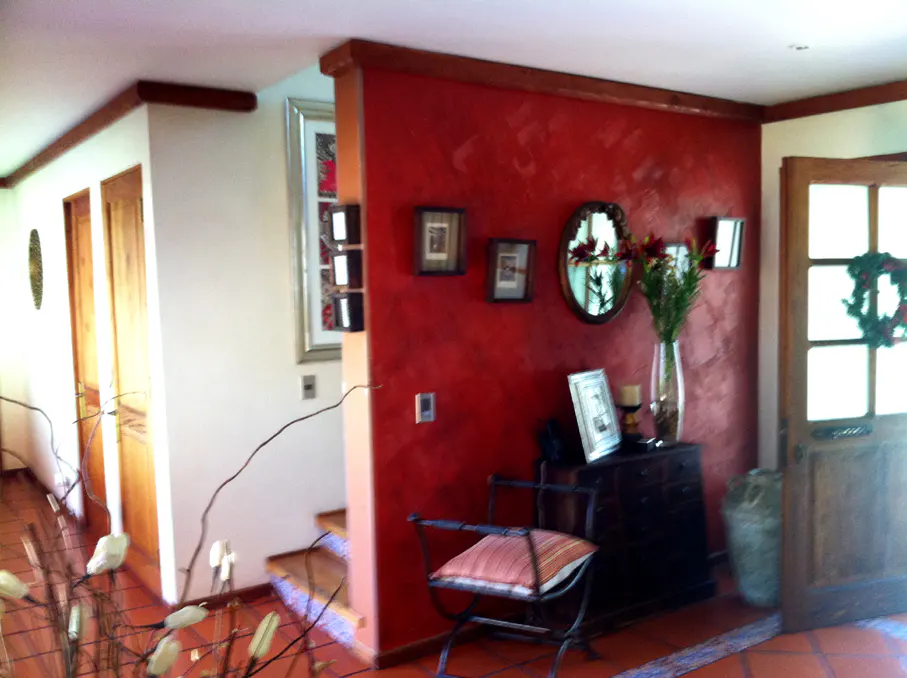 Interior view of a home with red accent wall, dark wood trim, and tile flooring. A chair sits near a dark wood cabinet with flowers and framed photos. A door with a wreath is on the right.