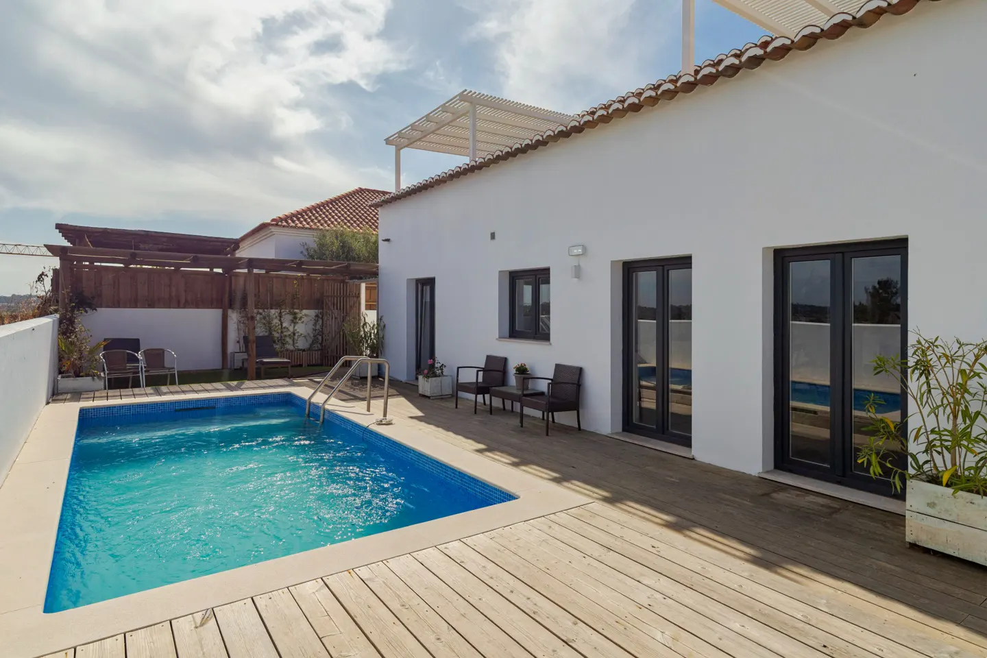 Outdoor pool area with blue tiled pool, wooden deck, white building, black framed windows, and patio furniture.