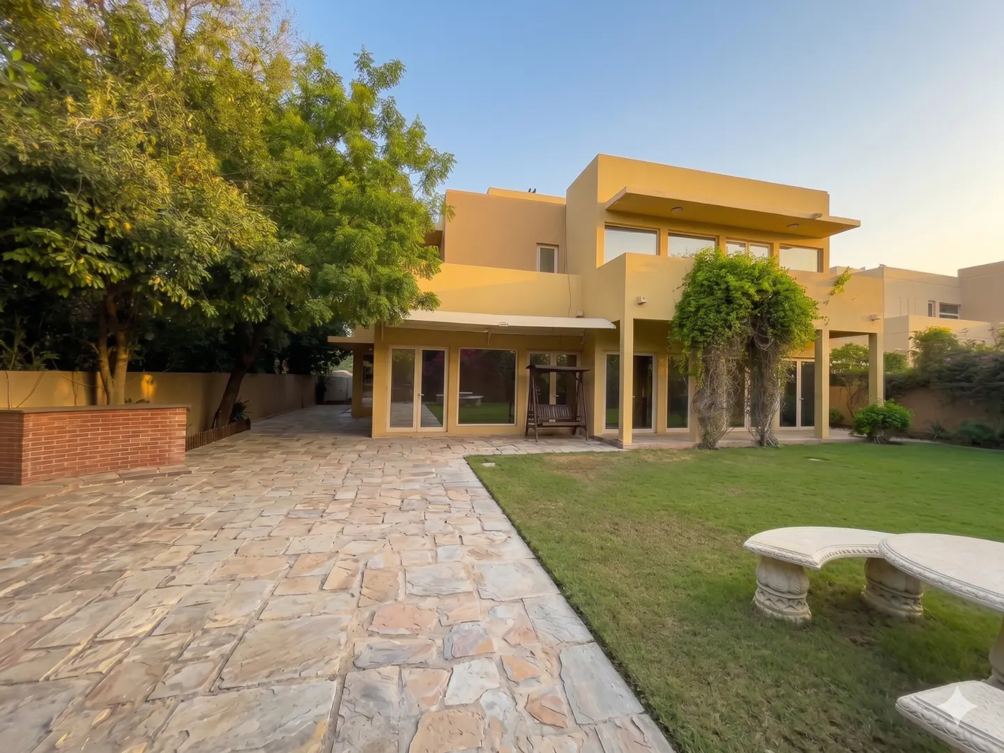 Tan two-story house with a stone patio, green lawn, and trees under a blue sky.