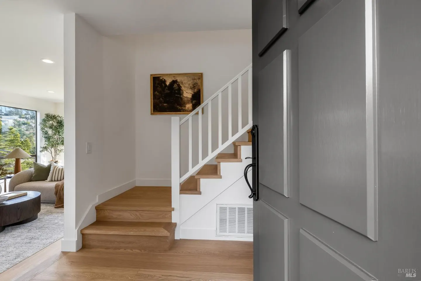 Open gray front door reveals a bright foyer with wood floors, stairs with white railings, and a glimpse of the living room.