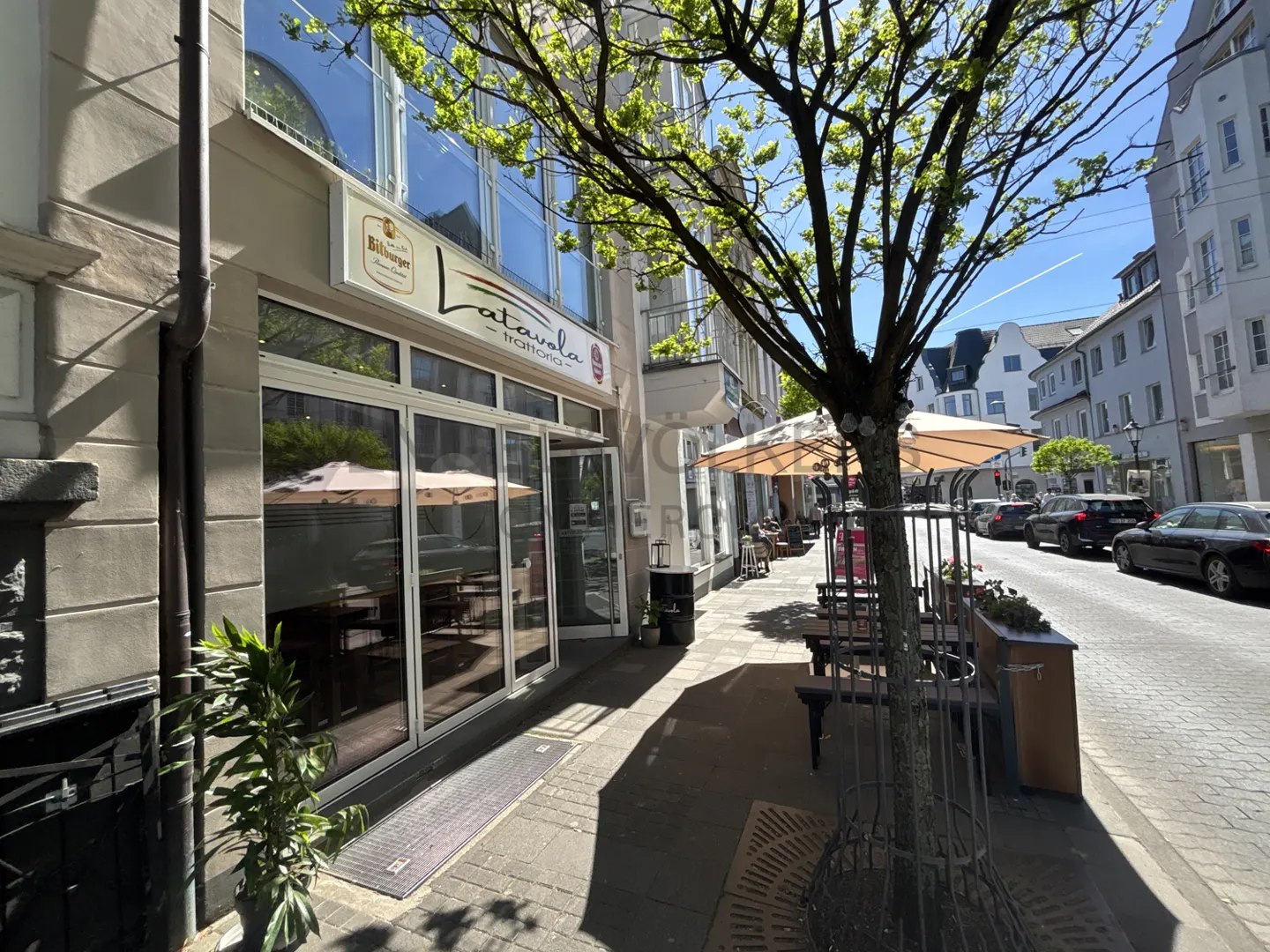 Exterior view of La Tavola Trattoria with outdoor seating under beige umbrellas on a sunny day. Cars are parked along the street.