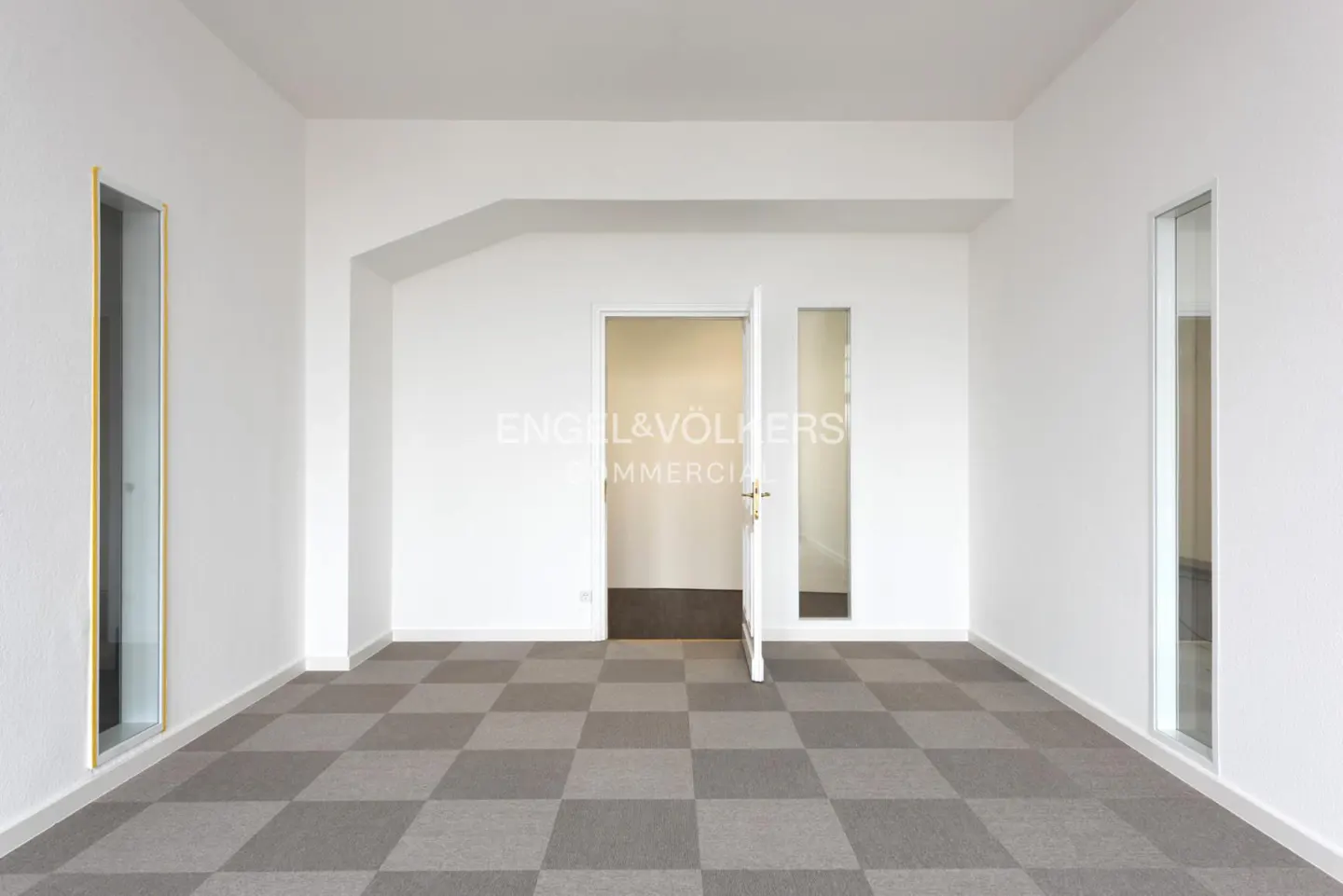 Empty office space with white walls, gray checkered carpet, and an open door leading to another room. Windows on either side of the room.