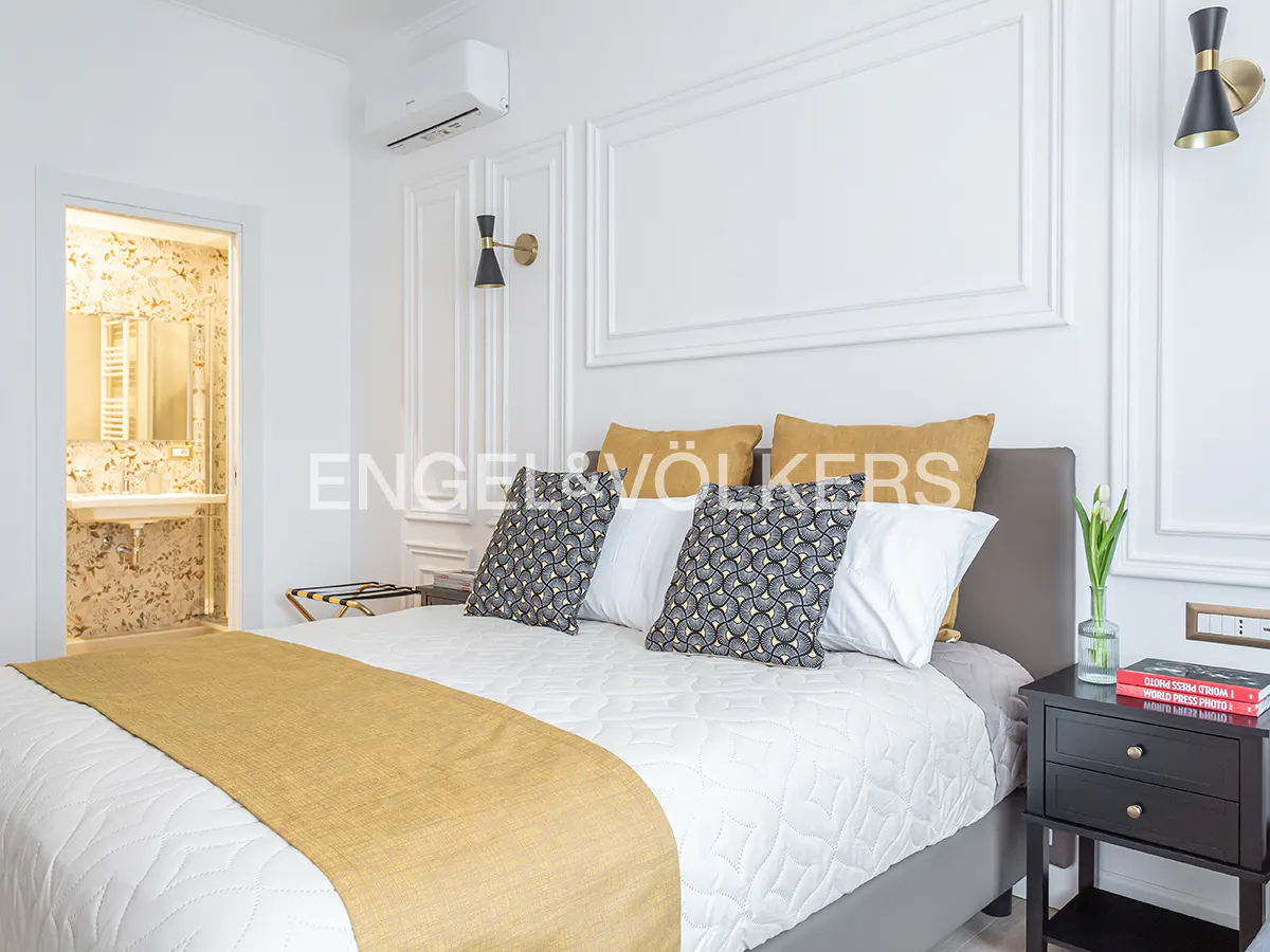 Bedroom with a white quilted bedspread, gold runner, patterned pillows, and a view into a floral-wallpapered bathroom.