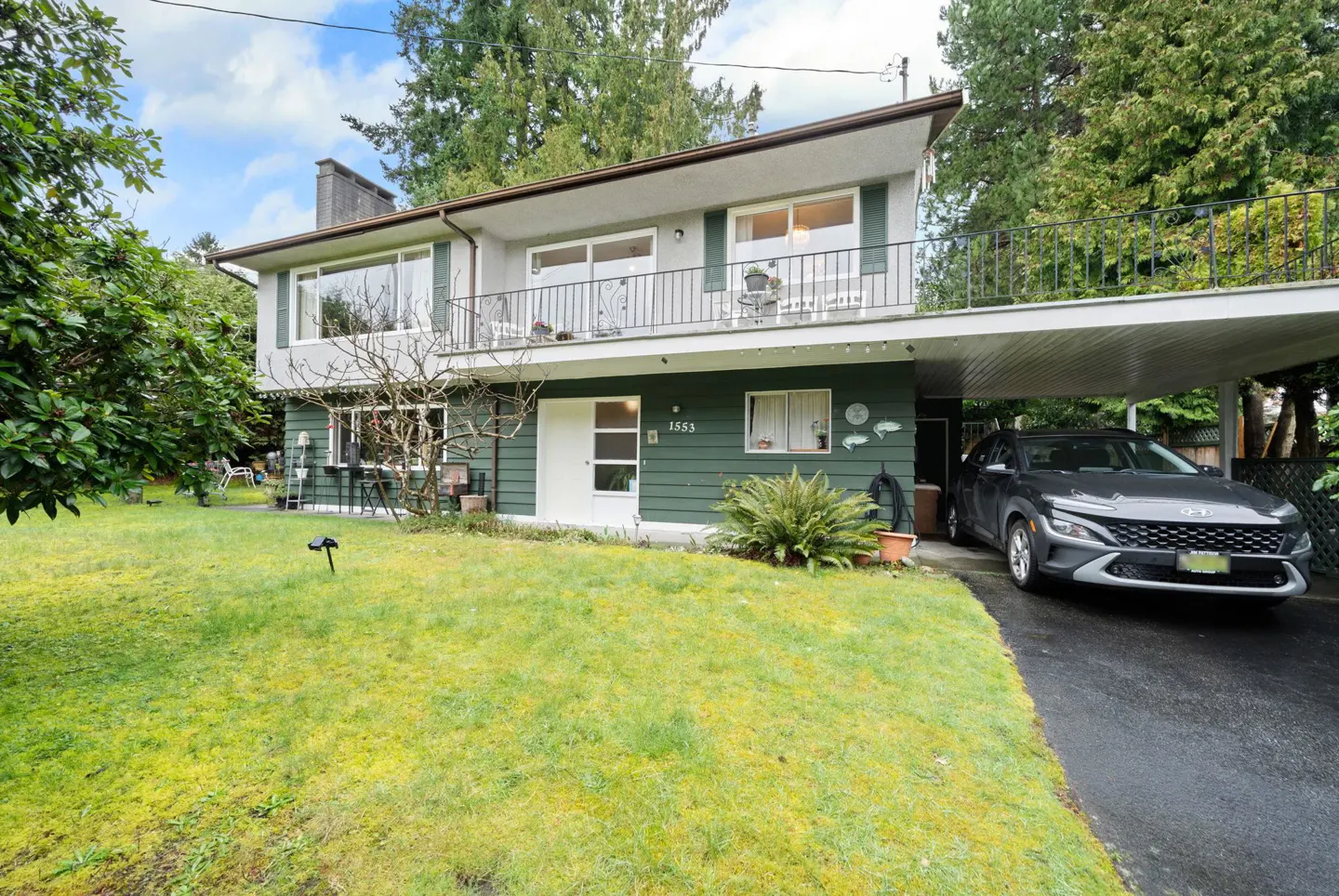 Two-story home with green siding, a balcony, and a carport with a gray car parked underneath. A green lawn is in front.