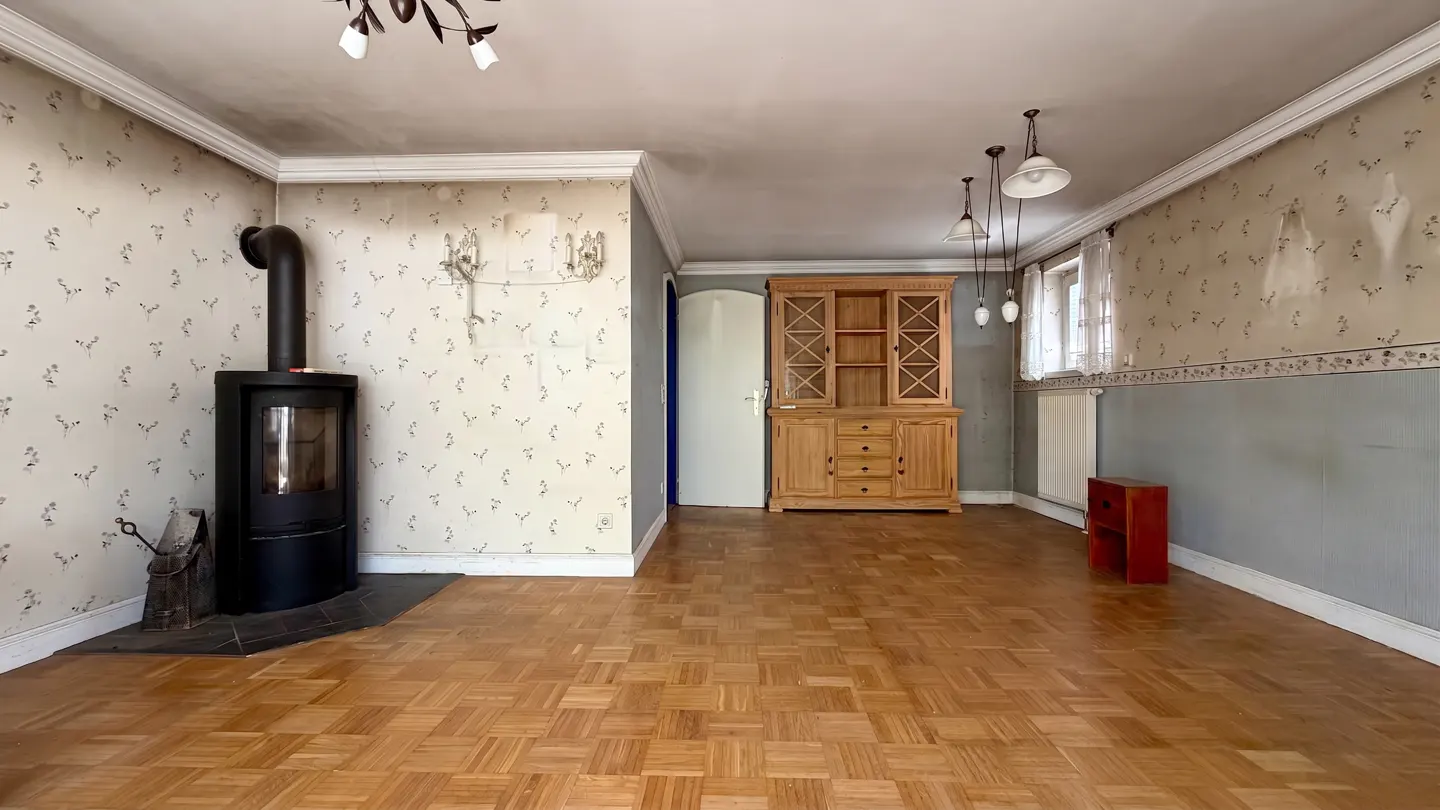 A living room with parquet floors, floral wallpaper, and a black wood-burning stove. A wooden hutch stands against the wall.