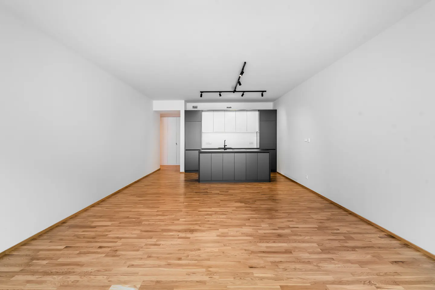 An empty room with hardwood floors, white walls, and a gray and white kitchen in the background.