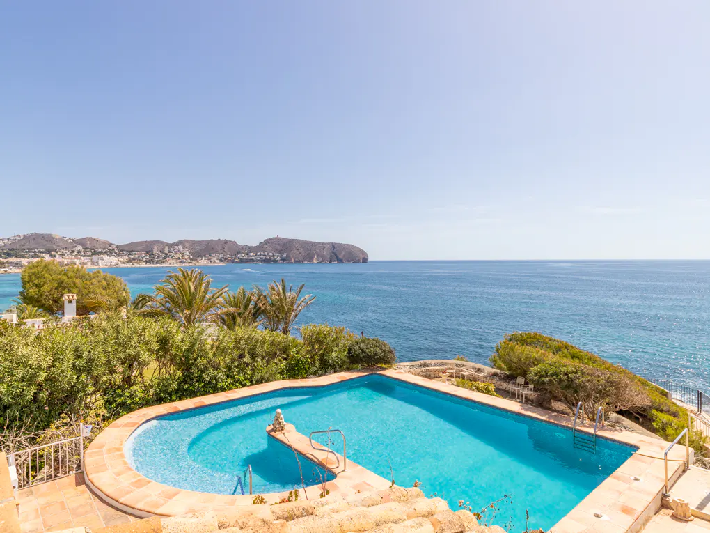 A turquoise pool overlooks the ocean on a sunny day. Palm trees and greenery surround the pool area.