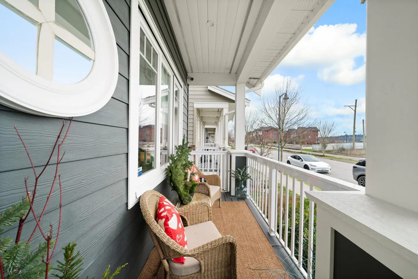A cozy porch with wicker chairs, red heart pillows, and a brown rug. White railings offer street views with cars and buildings. Gray siding and white trim complete the exterior.