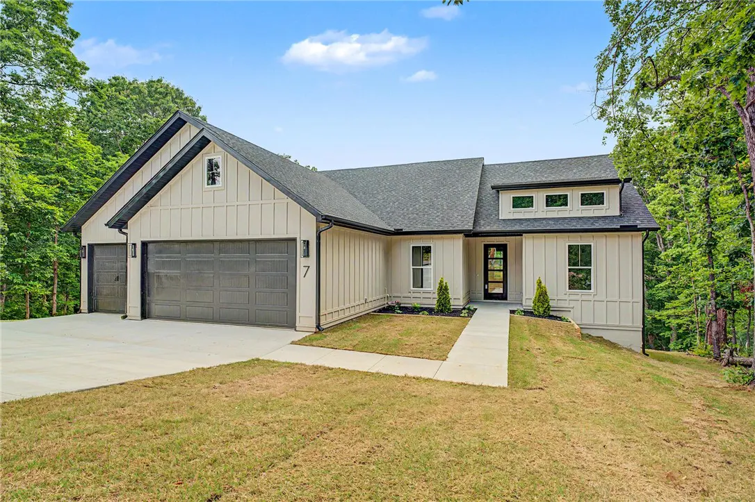 A modern, single-story home with light beige siding, a gray roof, and a two-car garage, surrounded by green trees.
