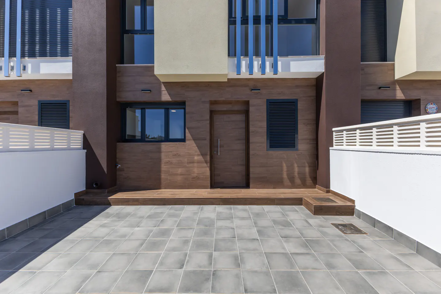 Exterior view of a modern brown and beige townhouse with a gray tiled patio and a brown front door.