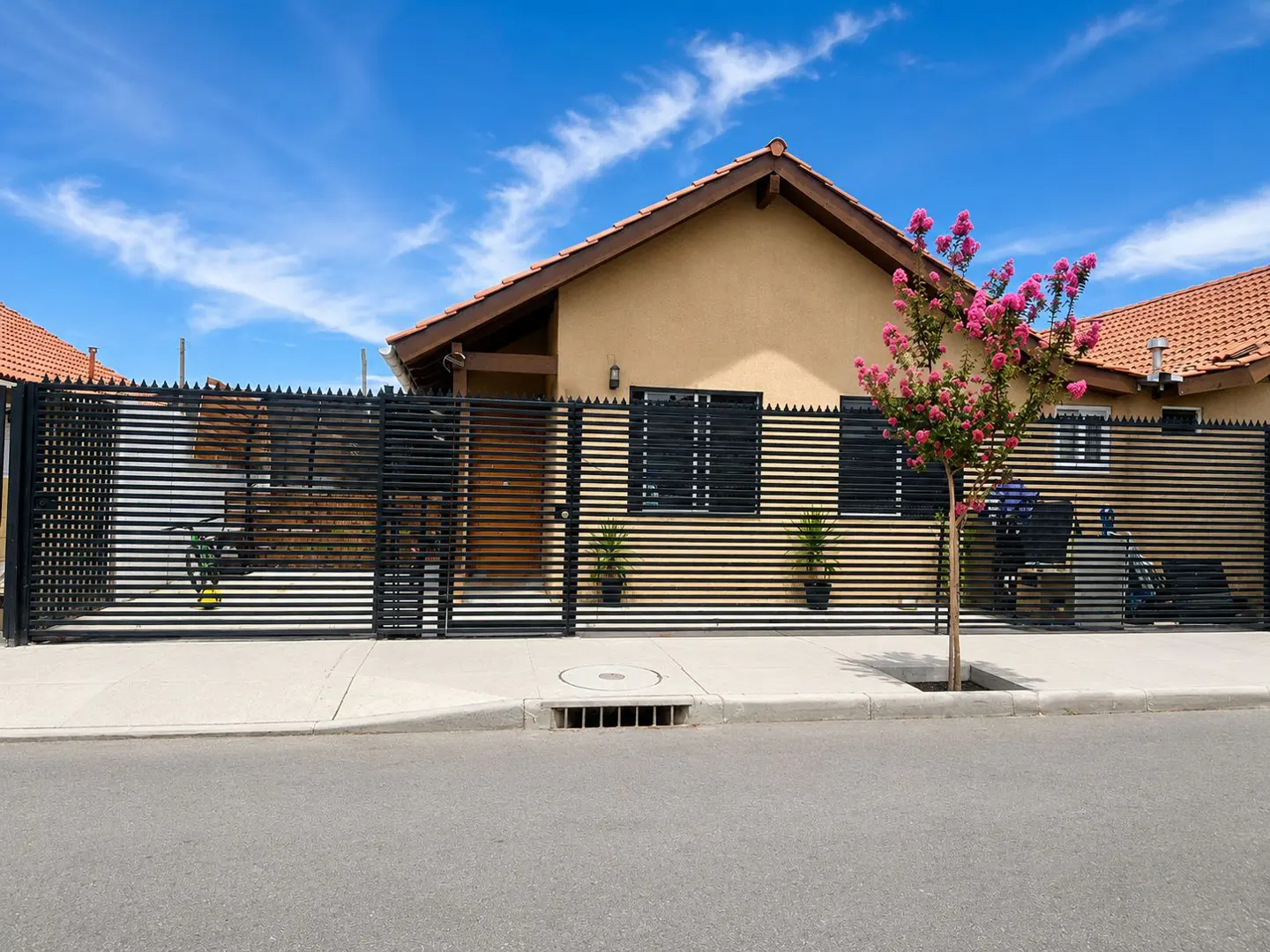 Tan house with a brown roof, black fence, and pink flowering tree on a sunny day.