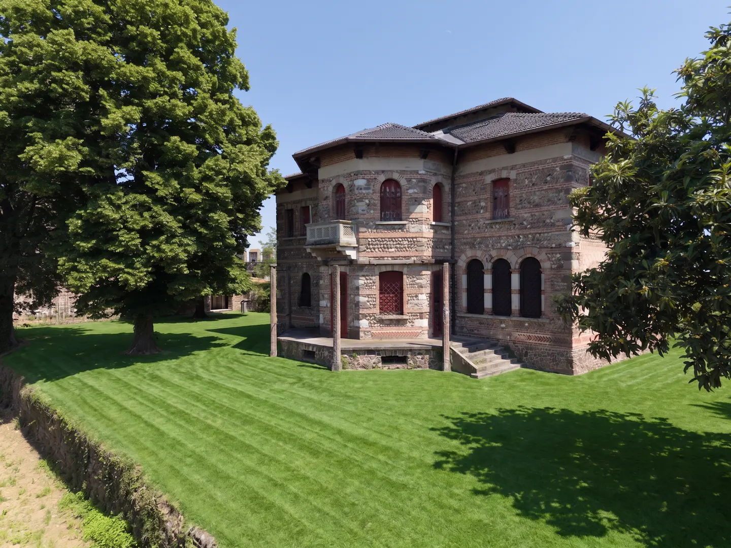 A historic stone villa with arched windows and a balcony, set in a large, manicured green lawn under a clear blue sky.
