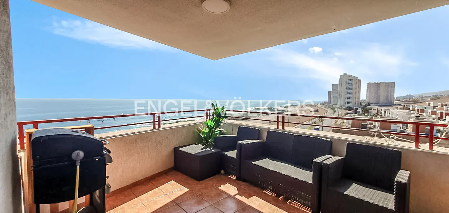Balcony with ocean view features black wicker furniture, grill, and red railing. Tall buildings are visible in the background.