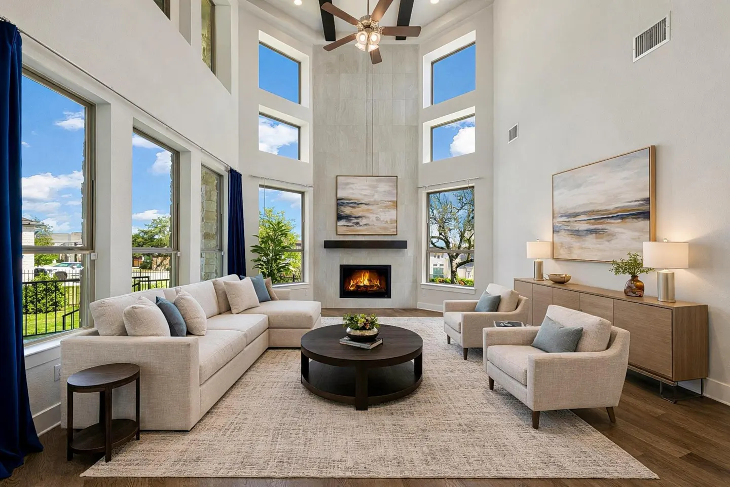 Bright living room with high ceilings, large windows, and a fireplace. Beige sectional sofa, two armchairs, and a round coffee table on a patterned rug.