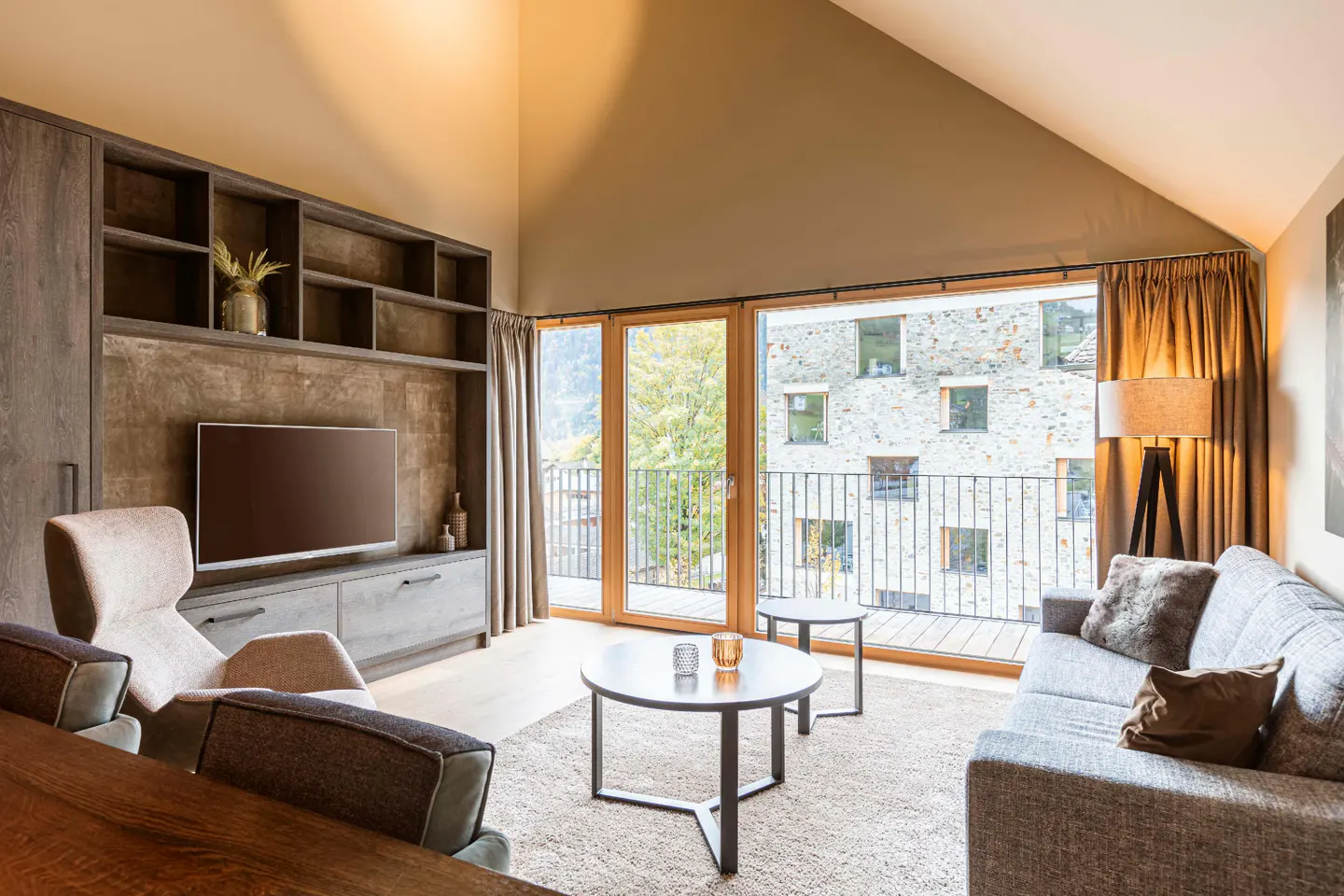 Modern living room with vaulted ceiling, large windows, grey sofa, armchair, and built-in shelving unit with a TV.