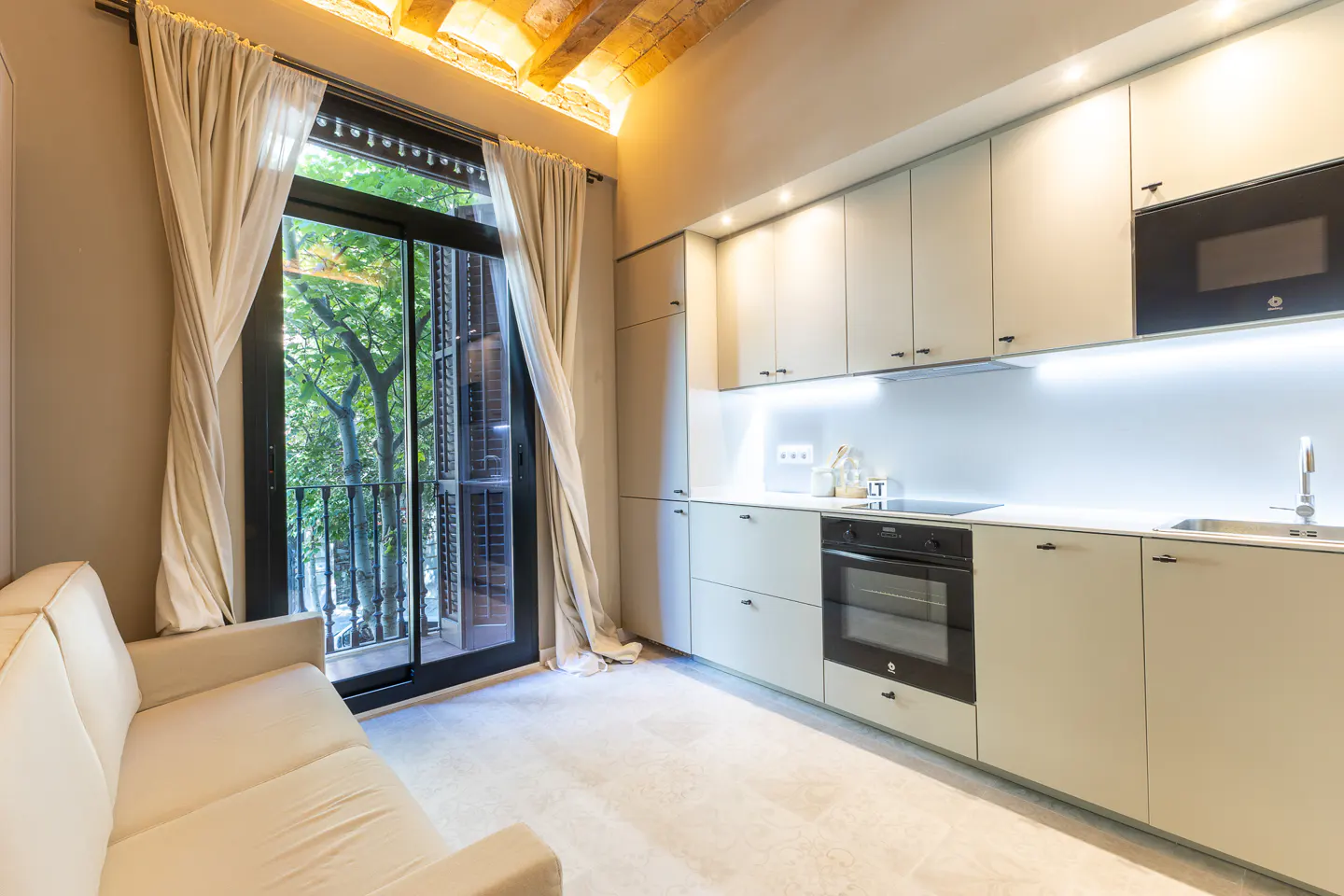 A modern kitchen with light green cabinets, black appliances, and a cream sofa next to a balcony door with white curtains.