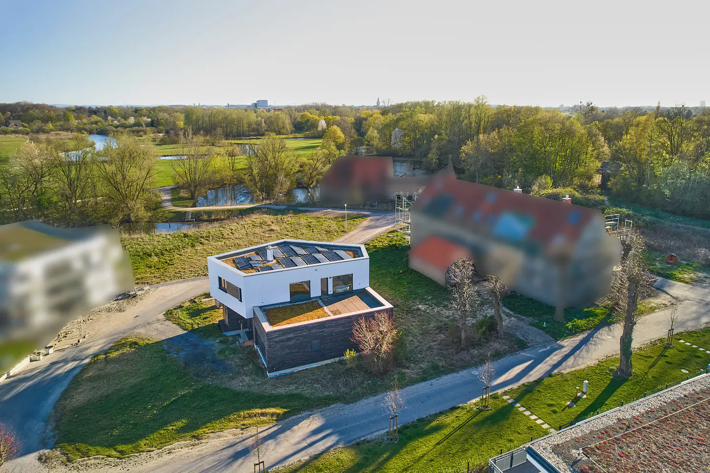 Aerial view of a modern white house with solar panels and a green roof, set in a lush green landscape with trees and a river.