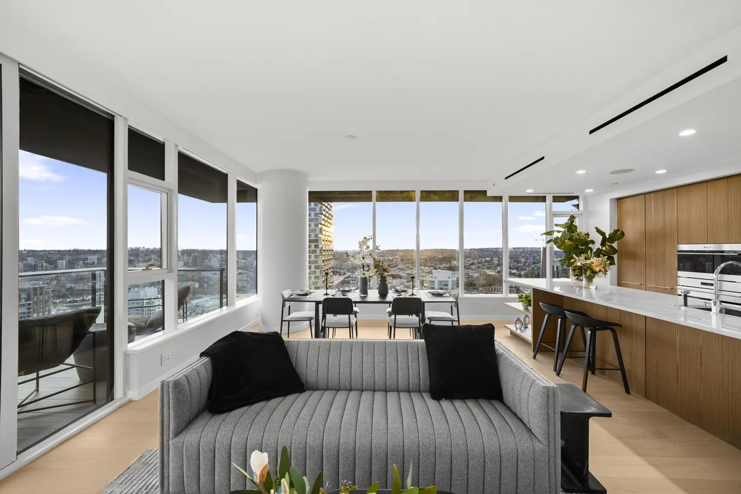 Bright, modern condo interior with a gray sofa, dining table, and kitchen island, all with a city view through large windows.