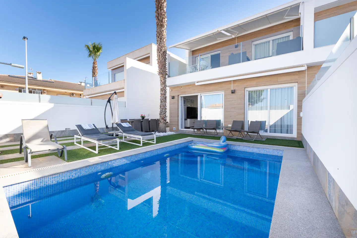 Modern villa backyard with a blue swimming pool, lounge chairs on artificial grass, and a tall palm tree under a clear sky.