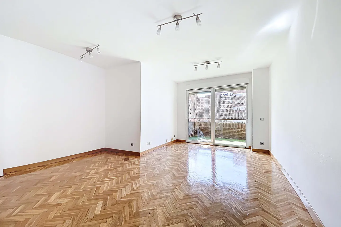 Bright, empty room with white walls, herringbone wood floor, and sliding glass doors to a balcony. Track lighting on the ceiling.