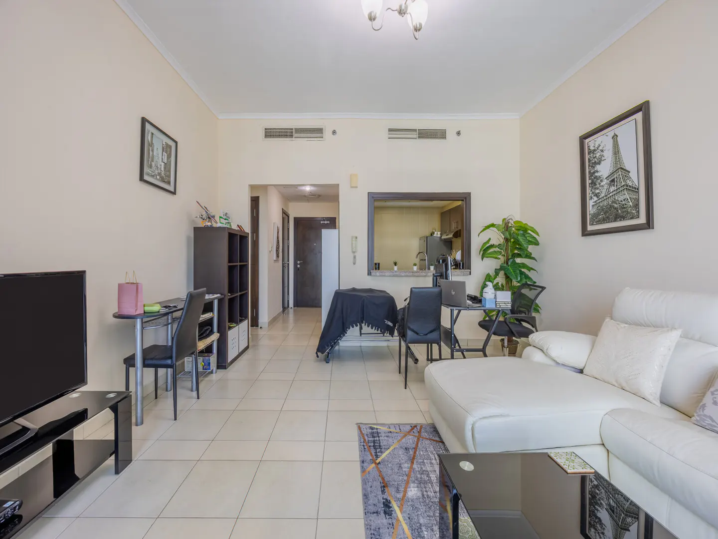 A bright living room with beige walls, tile floors, and a white sectional sofa. There is a black TV stand, desk, and chairs. A picture of the Eiffel Tower hangs on the wall.