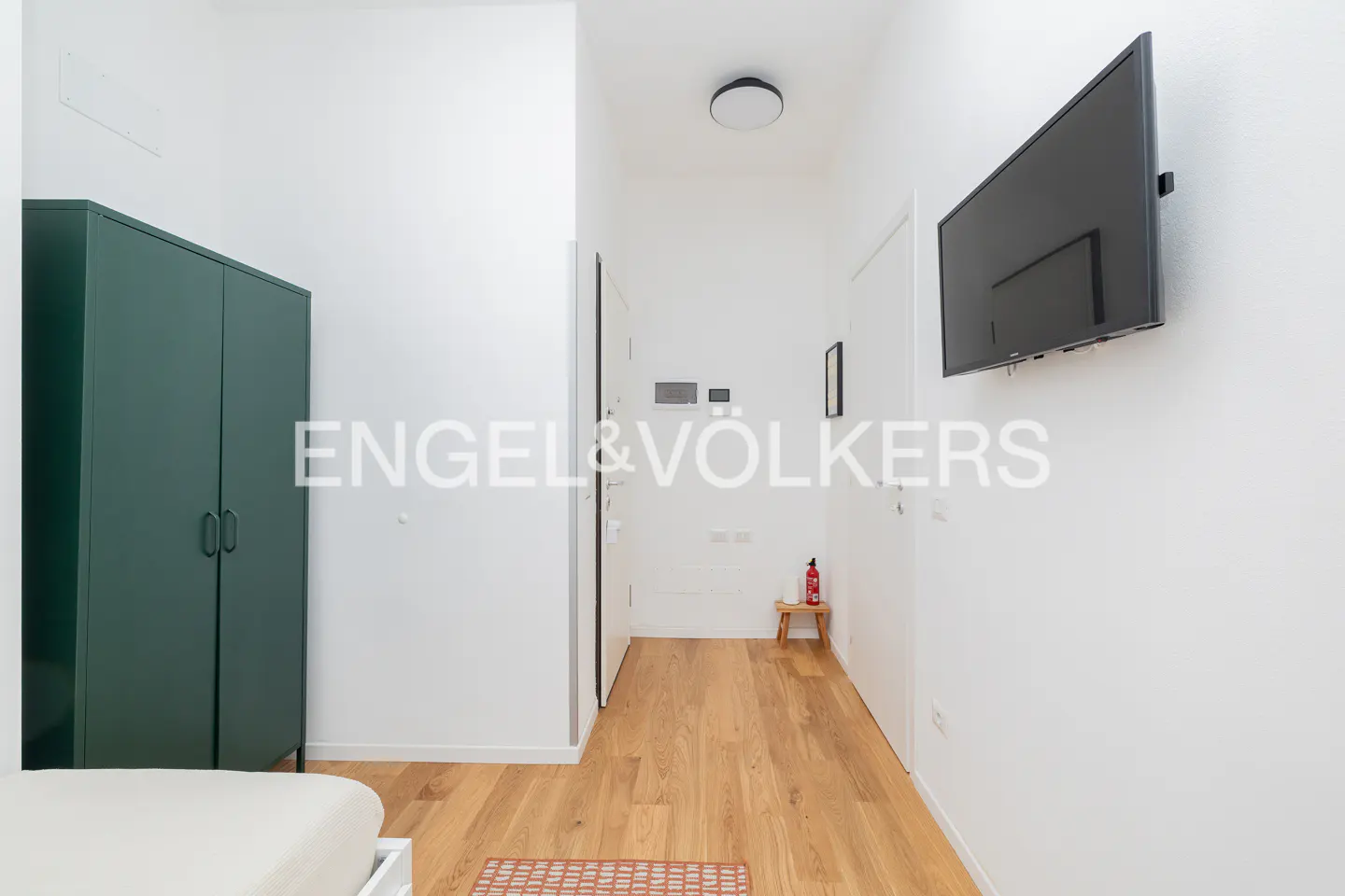 A bright hallway with light wood floors, a dark green wardrobe, a wall-mounted TV, and a small stool with a fire extinguisher.