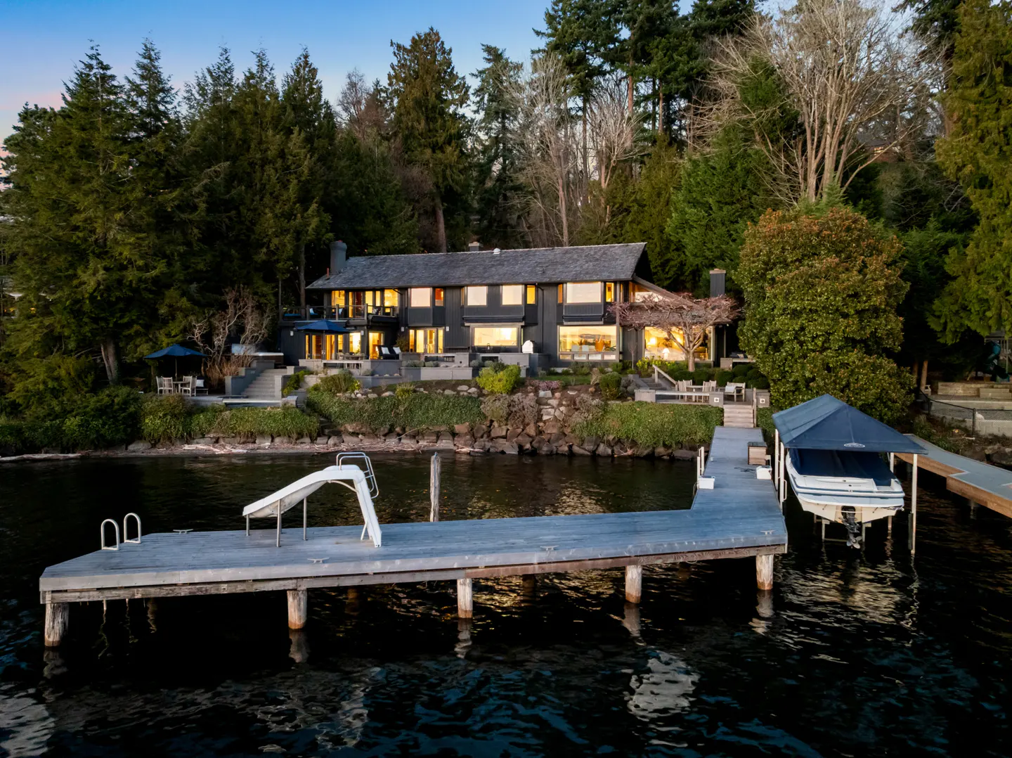 Lakefront home with a private dock. The dark gray house is surrounded by trees. The dock has a boat lift and a slide.