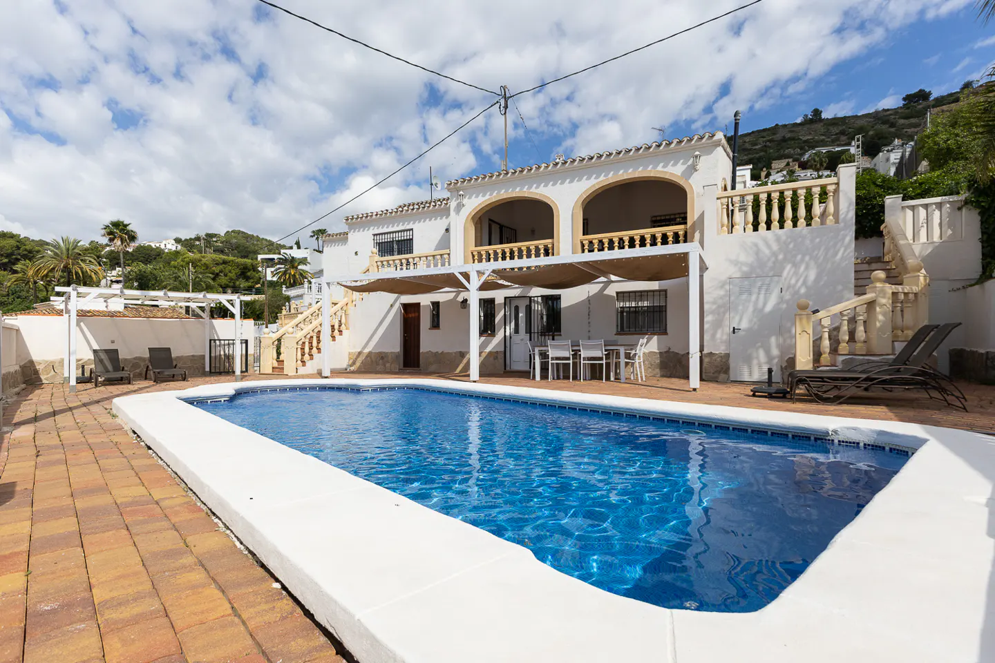 A white villa with a blue swimming pool, brick patio, and outdoor furniture on a sunny day.