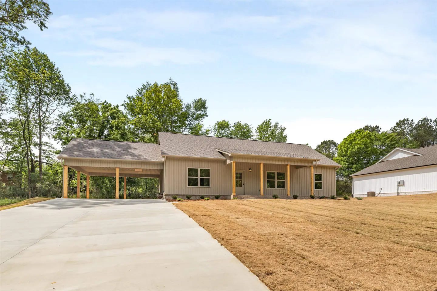 Tan house with a carport and a concrete driveway, surrounded by trees and dry grass.