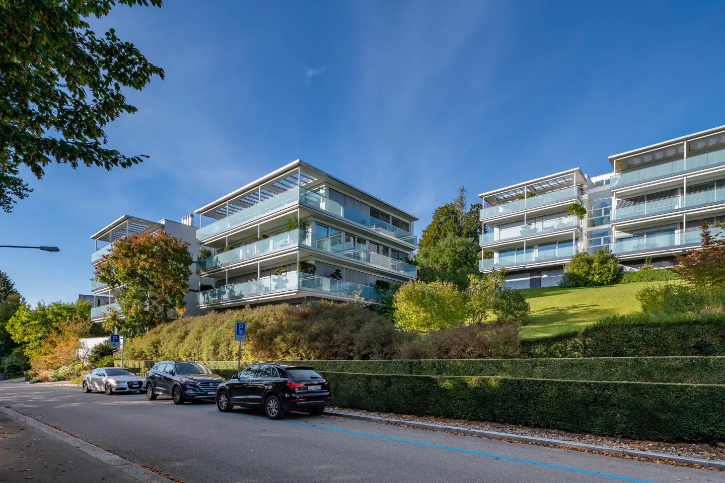 Modern white apartments with glass balconies sit atop a green hill. Cars are parked on the street below, under a clear blue sky.