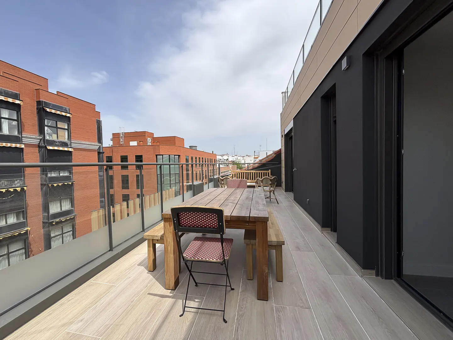 Outdoor patio with a wooden table, benches, and chair. A glass railing overlooks red brick buildings under a blue sky.