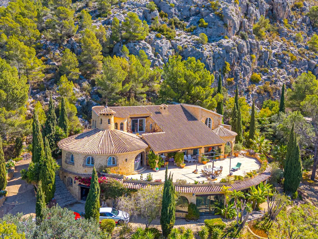 Aerial view of a tan stone house with a brown tile roof, surrounded by green trees and a rocky hillside. A patio with furniture is visible.